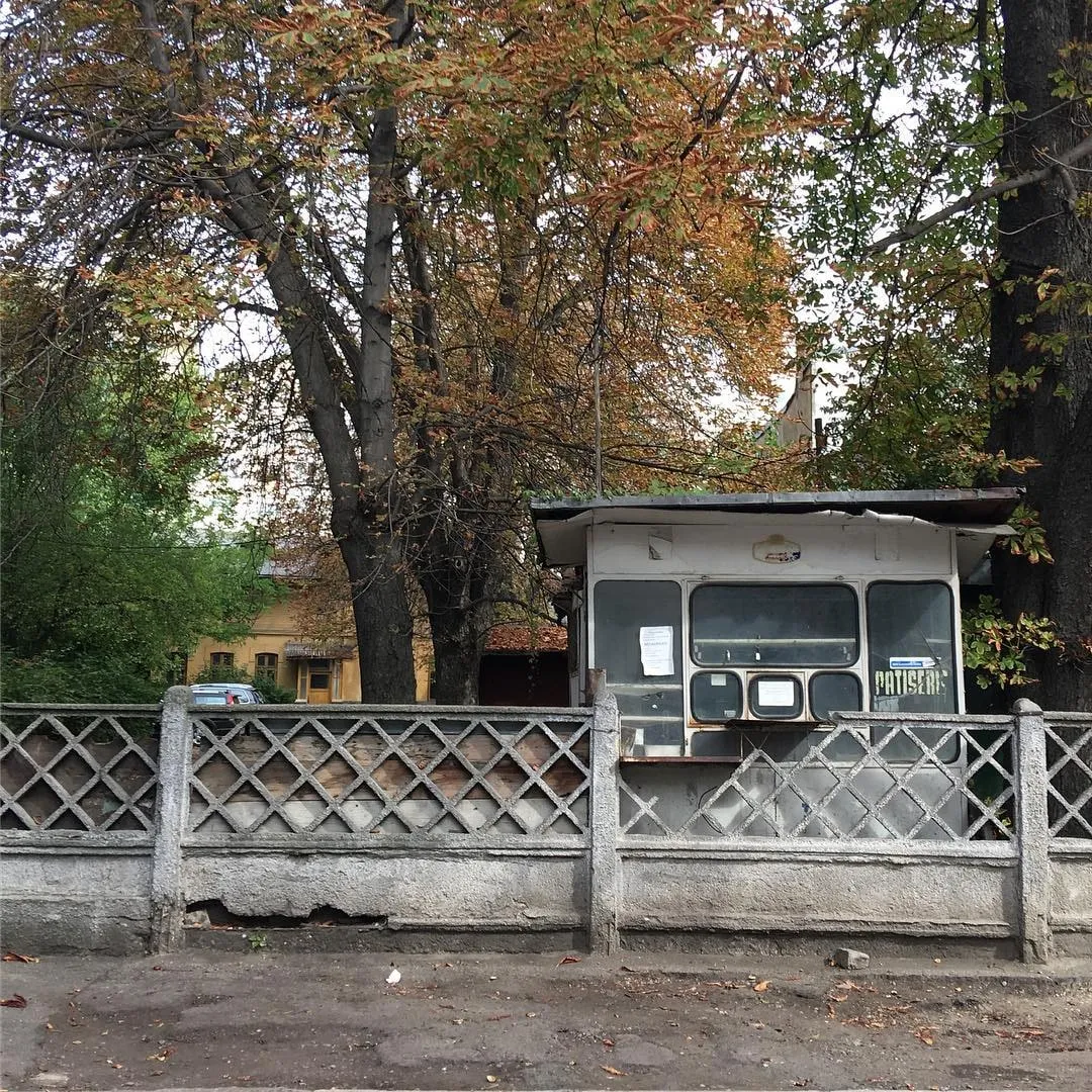 An old white kiosk marked PATISERIE stands behind a concrete lattice fence under autumn trees