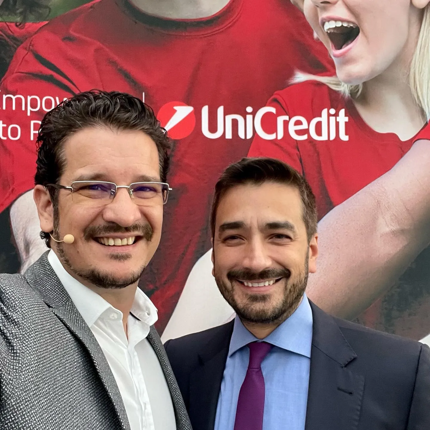 Sebastian Cochinescu și un alt bărbat zâmbesc în timp ce își fac un selfie în fața unui ecran mare UniCredit.