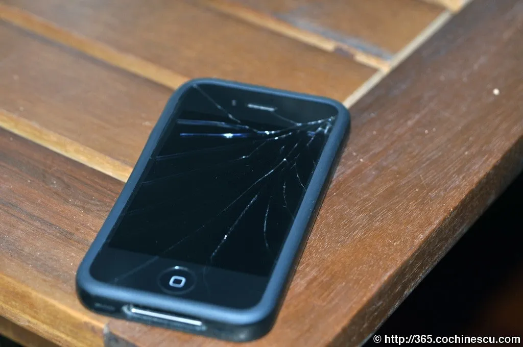 An iPhone 4 with a cracked screen is photographed on a wooden table.